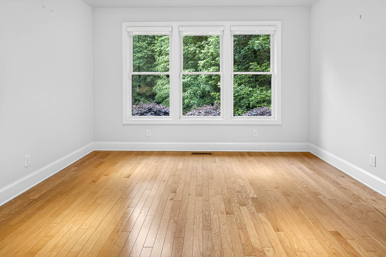 Empty room with light-colored hardwood floors and white walls, featuring three large windows that overlook green trees and shrubs outside. Natural light fills the space, creating a bright and airy atmosphere.