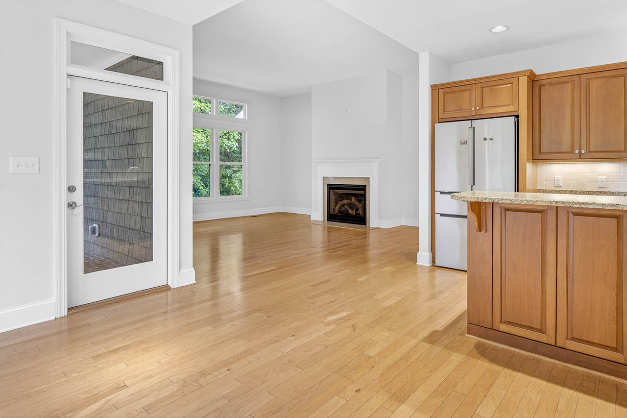 Spacious, empty room with light wood floors, a kitchen area with wooden cabinets and granite counters, a white fridge, a door with glass panes, large windows, and a white fireplace in the living area.