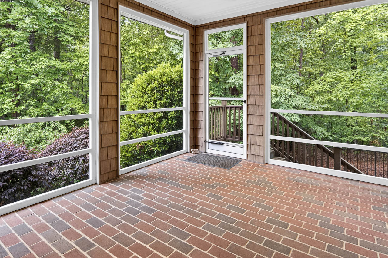 A screened-in porch with brown shingled walls, red brick flooring, and large windows overlooking lush green trees; a door leads to wooden steps outside.