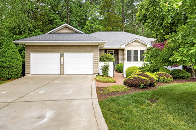 Single-story house with gray shingle roof, two-car garage, beige siding, white trim, and a landscaped front yard with green shrubs and trees. A wide driveway leads to the garage.