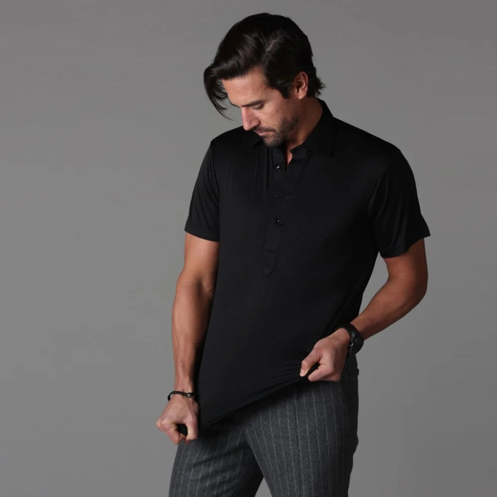 A man wearing the COLLARS AND CO - SEMI SPREAD COLLAR ORIGINAL POLO IN BLACK by COLLARS AND CO, paired with gray pinstripe pants, looks down while holding the hem of his shirt against a plain gray background.