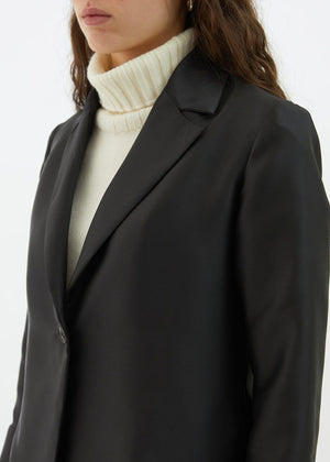 A person with long, wavy hair is wearing the BLACK BLAZER / JACKET by ALYKI LABORATORIO, made in Italy, over a white turtleneck sweater. They are standing against a white background with their arms crossed and have a serious expression on their face.