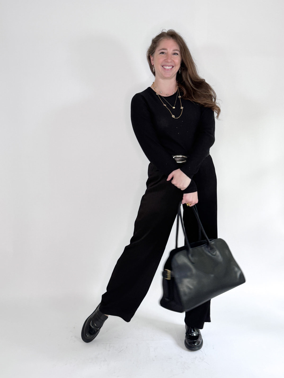 A woman smiles against a white backdrop, wearing a black top and ROSE AND CROWN - SATIN RIBBED PANT. She accessorizes with layered necklaces and a large black handbag, posing with one leg slightly raised for a relaxed look.