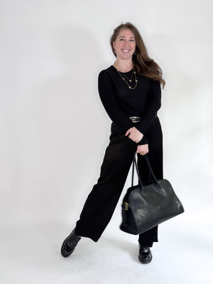 A woman smiles against a white backdrop, wearing a black top and ROSE AND CROWN - SATIN RIBBED PANT. She accessorizes with layered necklaces and a large black handbag, posing with one leg slightly raised for a relaxed look.