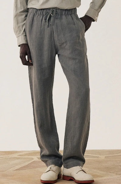 A person wearing HARTFORD - LINEN JOGGY PANTS IN ARMY with hands in pockets, a light shirt, and white dress shoes with brown soles stands on a patterned floor.