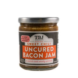 A jar of TBJ GOURMET Sweet Chili Uncured Bacon Jam sports a sleek black and orange label with the phrase "a warm and smoky spread" on the front. Sealed with a metal lid, this gluten-free jam is ideal for culinary adventures.