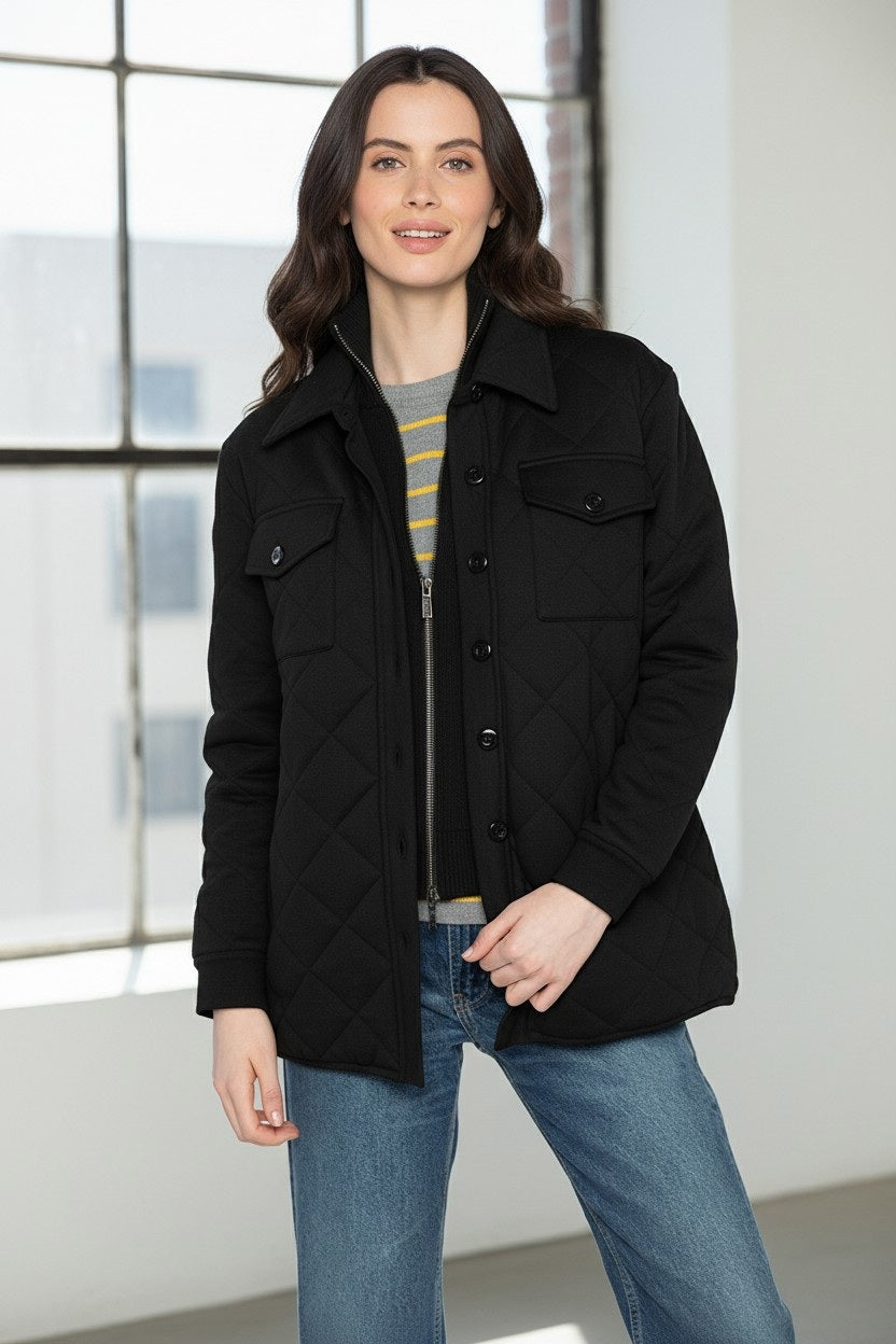 A woman stands indoors by a window, smiling softly as she touches her KINROSS CASHMERE TWO IN ONE QUILTED SHACKET, styled over a striped sweater and blue jeans.