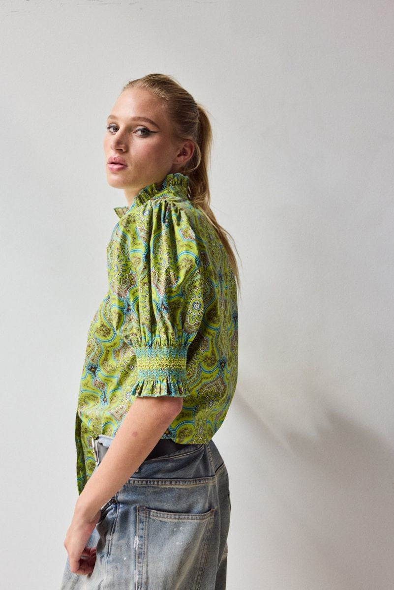 A woman with blonde hair in a ponytail wears the HOPE FOR FLOWERS Short Sleeve Smocked Shirt in Green Foulard, made of organic cotton, paired with faded blue jeans, standing with her back to the camera against a plain light background.