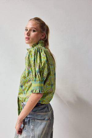 A woman with blonde hair in a ponytail wears the HOPE FOR FLOWERS Short Sleeve Smocked Shirt in Green Foulard, made of organic cotton, paired with faded blue jeans, standing with her back to the camera against a plain light background.