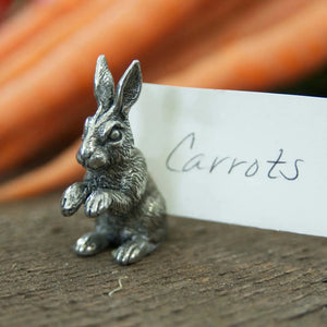 The VAGABOND HOUSE PEWTER RABBIT PLACE CARD HOLDER, detailed with intricate fur, sits elegantly on wood next to a white "Carrots" card. Blurry orange carrots in the background add rustic charm.