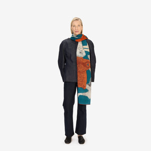 A woman with short blonde hair wears a dark jacket and the INOUI EDITIONS TOUTOU Dogs wool scarf in duck blue from INOUI, featuring playful animal illustrations in orange, teal, and cream, standing against a plain white background.