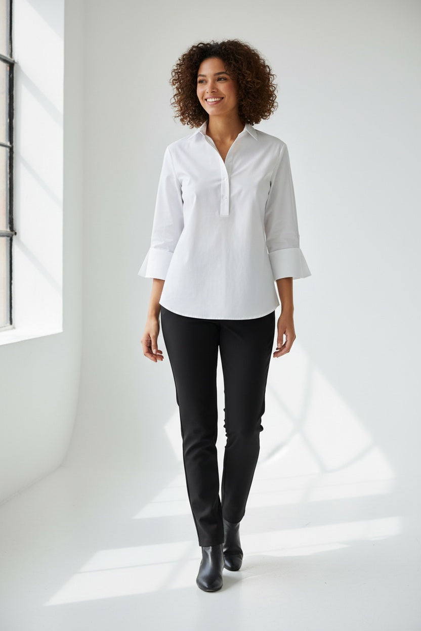 A woman with short dark hair models the HINSON WU - AILEEN 3/4 SLEEVE POLISHED COTTON SHIRT in navy, featuring a collared neckline, stretch fabric, and three buttons. She pairs it with white pants and poses against a plain light gray background.