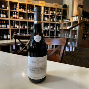 A bottle of ALBERT BICHOT ‘PASSETOUTGRAIN’ 2021 red wine stands on a white table in front of a wine rack.