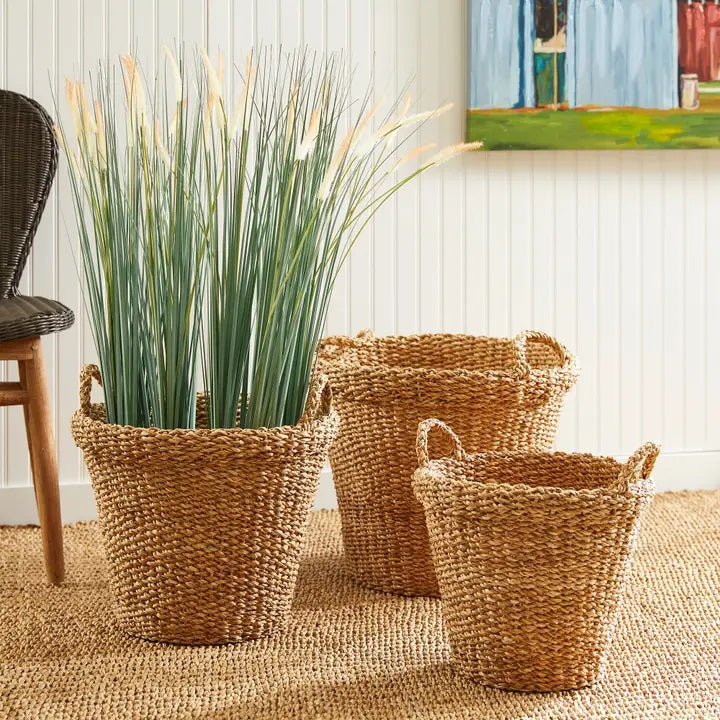 The NAPA HOME AND GARDEN Seagrass Tapered Basket with Handles and Cuff, Small, sits on a textured rug before a white paneled wall, alongside two other baskets, with part of a chair and a colorful painting in the background.
