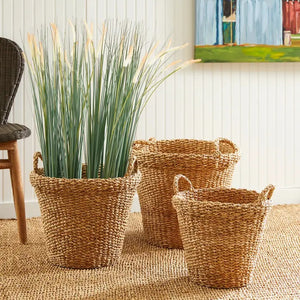 A NAPA HOME AND GARDEN Seagrass Tapered Basket with Handles and Cuff (Medium) sits on a woven rug, holding tall decorative green grass, with a chair and colorful painting in the background.