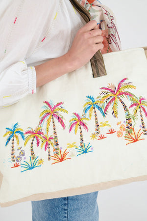 A person in a white shirt and blue jeans holds the STORIATIPIC BEACH TOTE BAIA, an embroidered cotton bag featuring colorful palm trees and plants.