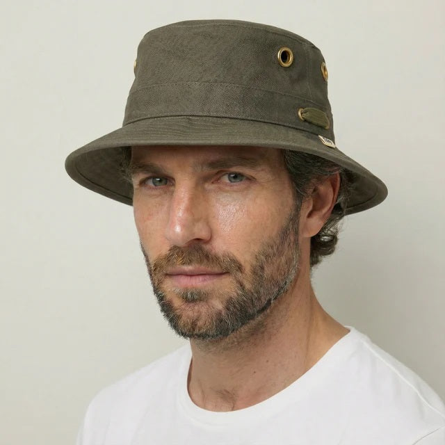 A person wearing the TILLEY - ICONIC T1 BUCKET HAT IN OLIVE with yellow eyelets and a white crew-neck shirt looks slightly to the side against a plain light background. The TILLEY hat offers UPF 50+ sun protection.