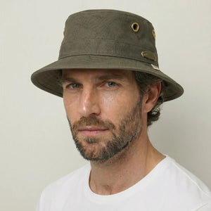 A person wearing the TILLEY - ICONIC T1 BUCKET HAT IN OLIVE with yellow eyelets and a white crew-neck shirt looks slightly to the side against a plain light background. The TILLEY hat offers UPF 50+ sun protection.