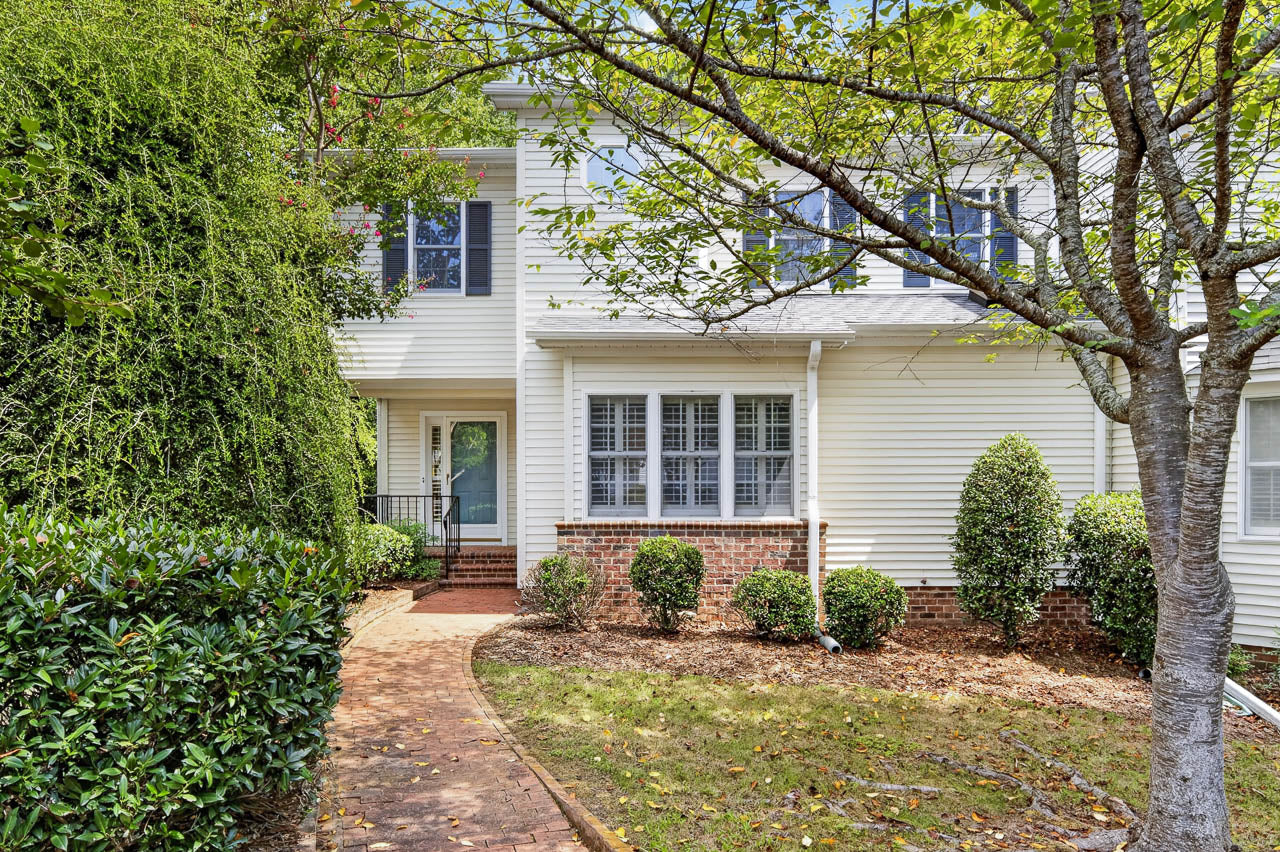 A white house with brick accents, a front porch, green shrubs, and a walkway leading to the door. A tree and lawn are in the foreground.