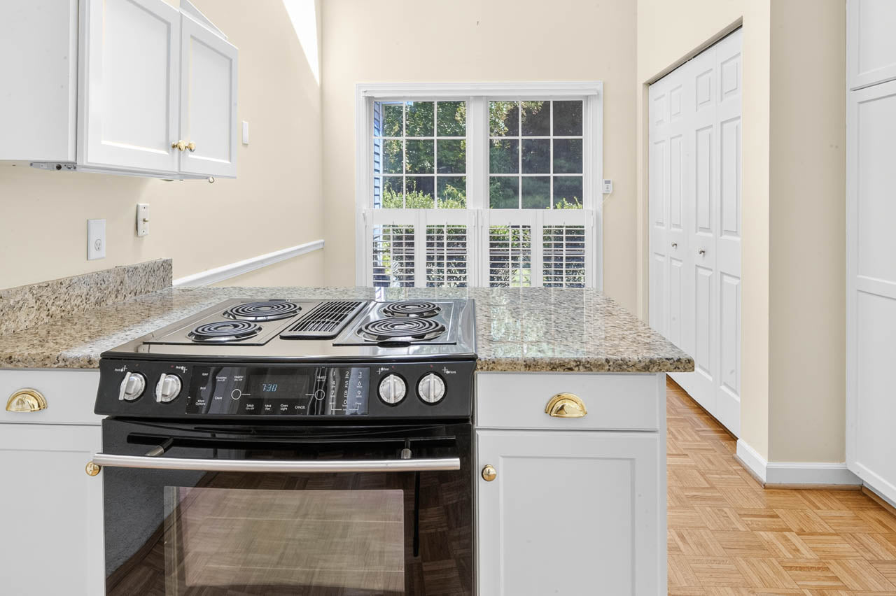 A modern kitchen with white cabinets, granite countertops, an electric stove, and oven. Sunlight streams in through large windows with white shutters, and a closet with double doors is visible.