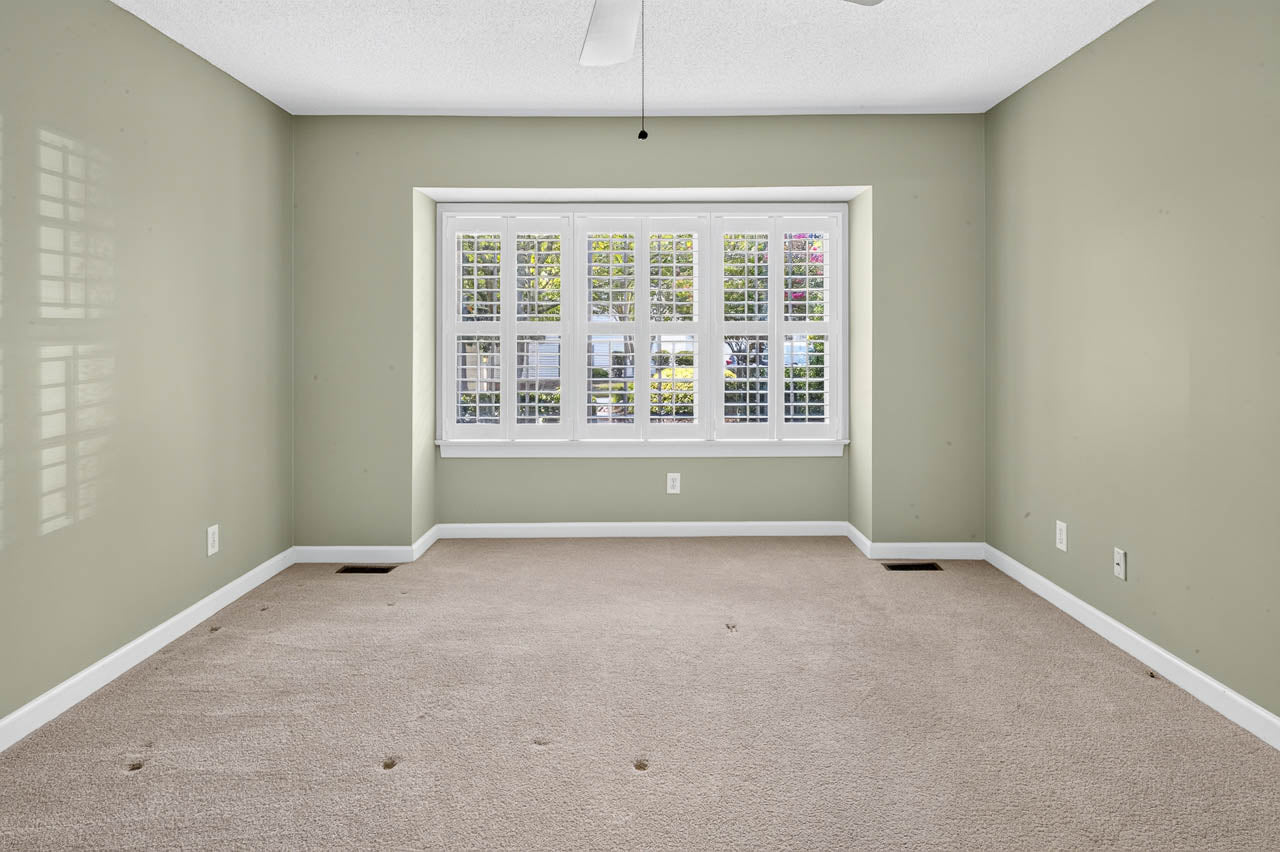 Empty room with light green walls, beige carpet, ceiling fan, and a large window with white shutters letting in natural light. There are small holes visible in the carpet.