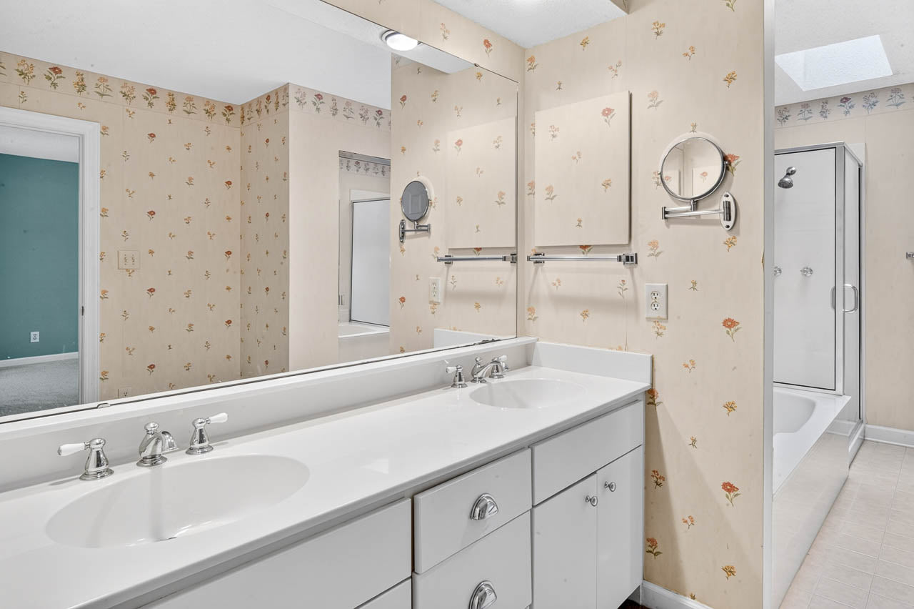 A bathroom with double sinks, a wide mirror, beige floral wallpaper, towel racks, and a glass shower. A doorway leads to a carpeted room with green walls.