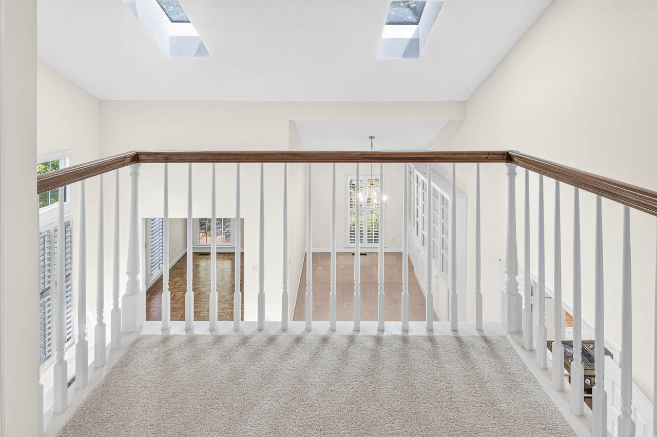 View from an upstairs landing with white railing, beige carpet, skylights in the ceiling, and a chandelier hanging over a spacious, open area below.
