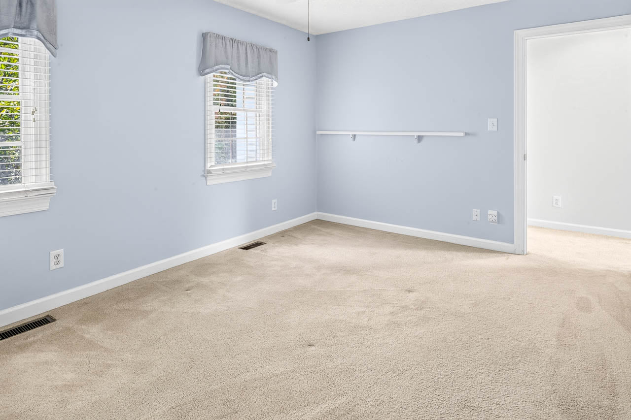 Empty room with light blue walls, beige carpet, two windows with white blinds and valances, and a small white shelf on the wall. Sunlight is coming in through the windows. Doorway leads to another room.