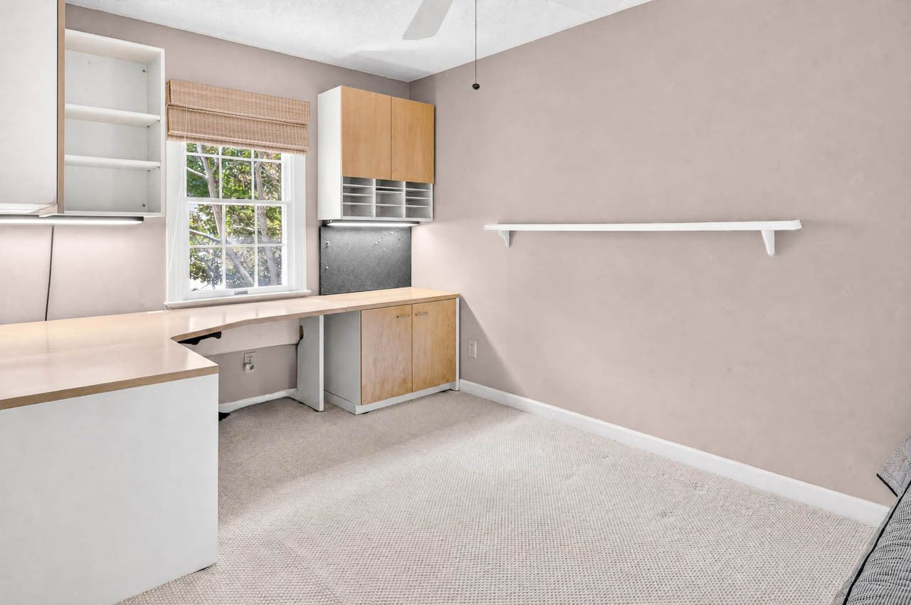 A home office with a built-in L-shaped desk, cabinets, shelves, a window with a woven shade, beige carpet, and light pink walls with a white wall shelf.
