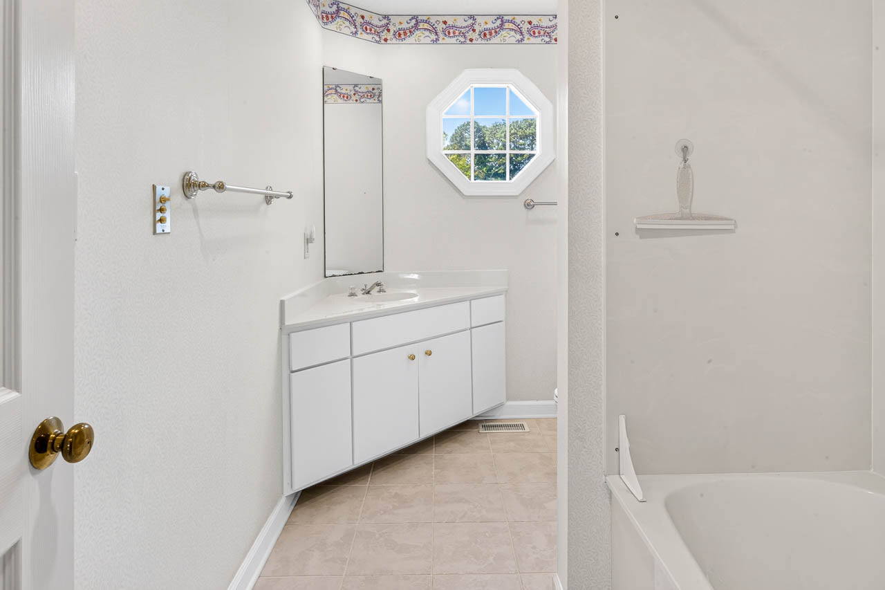 A bathroom with a white vanity, corner sink, mirror, octagonal window, beige tile floor, towel bars, and a white bathtub with a soap dish on the right.