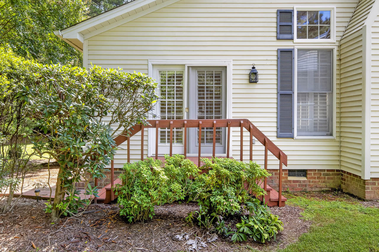Small wooden steps with a railing lead to a double glass door on a light yellow house, surrounded by green bushes and plants, with brick trim along the foundation.