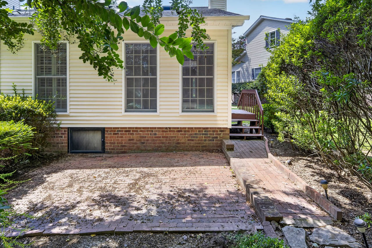 Brick patio area beside a yellow house with large windows, bordered by bushes and trees, and a small wooden deck with steps leading up to the house in the background.