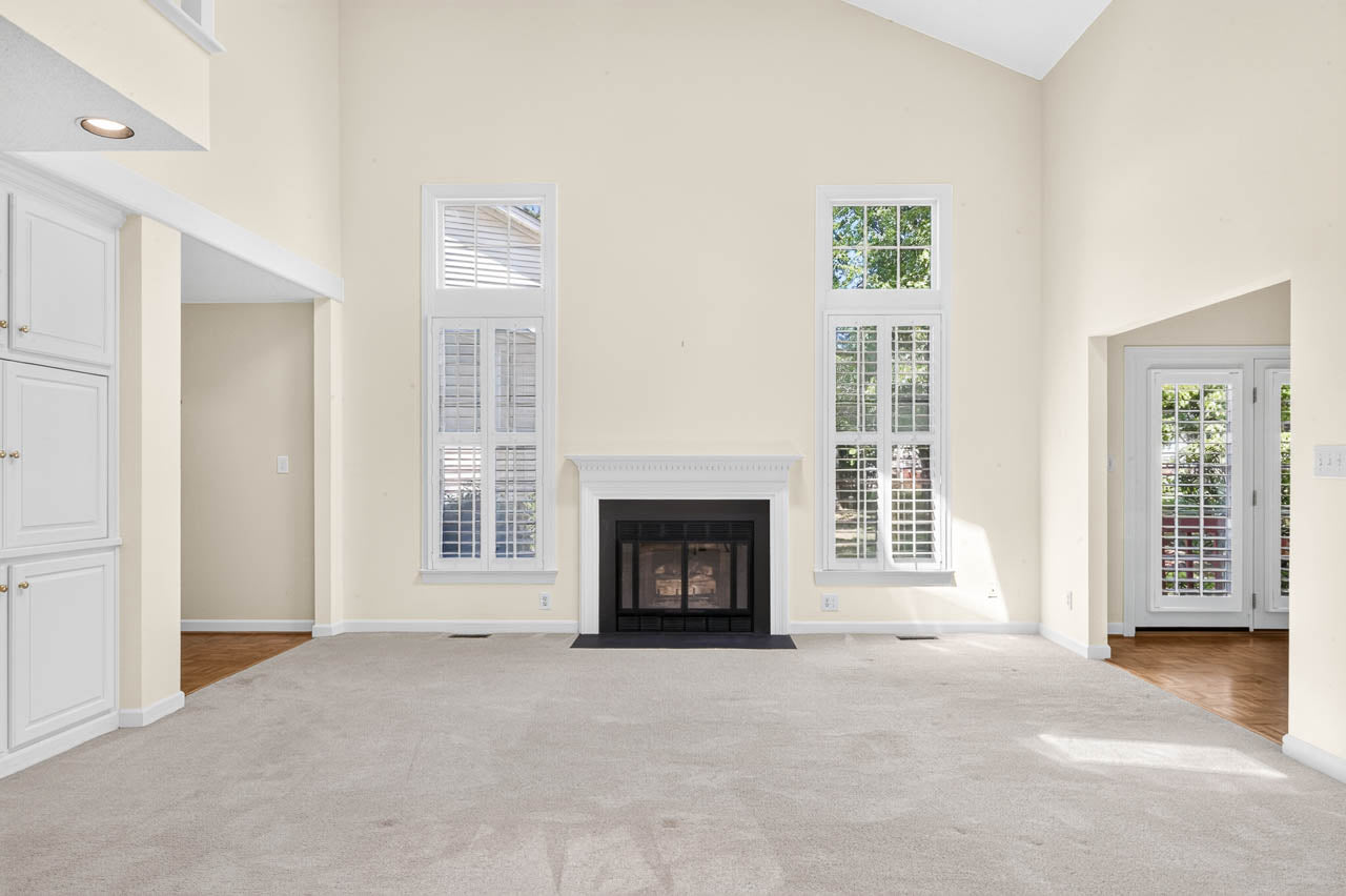 Bright living room with high ceilings, tall windows with white shutters, a white fireplace centered on one wall, cream-colored walls, and carpeted flooring. Sunlight streams through the windows and glass doors.