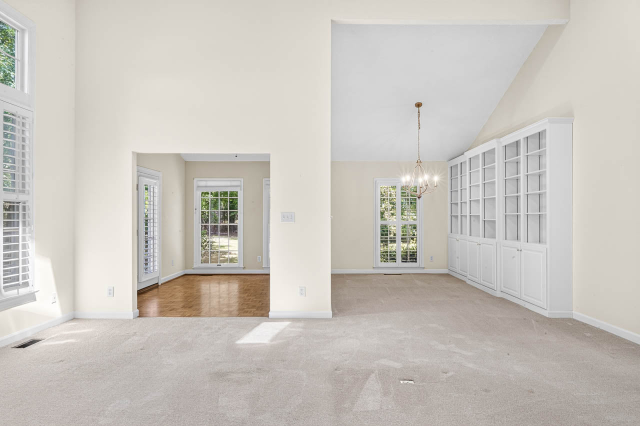 Bright, empty living and dining room with cream walls, carpeted floor, large windows, French doors, built-in shelving, chandelier, and hardwood flooring in the adjacent room.
