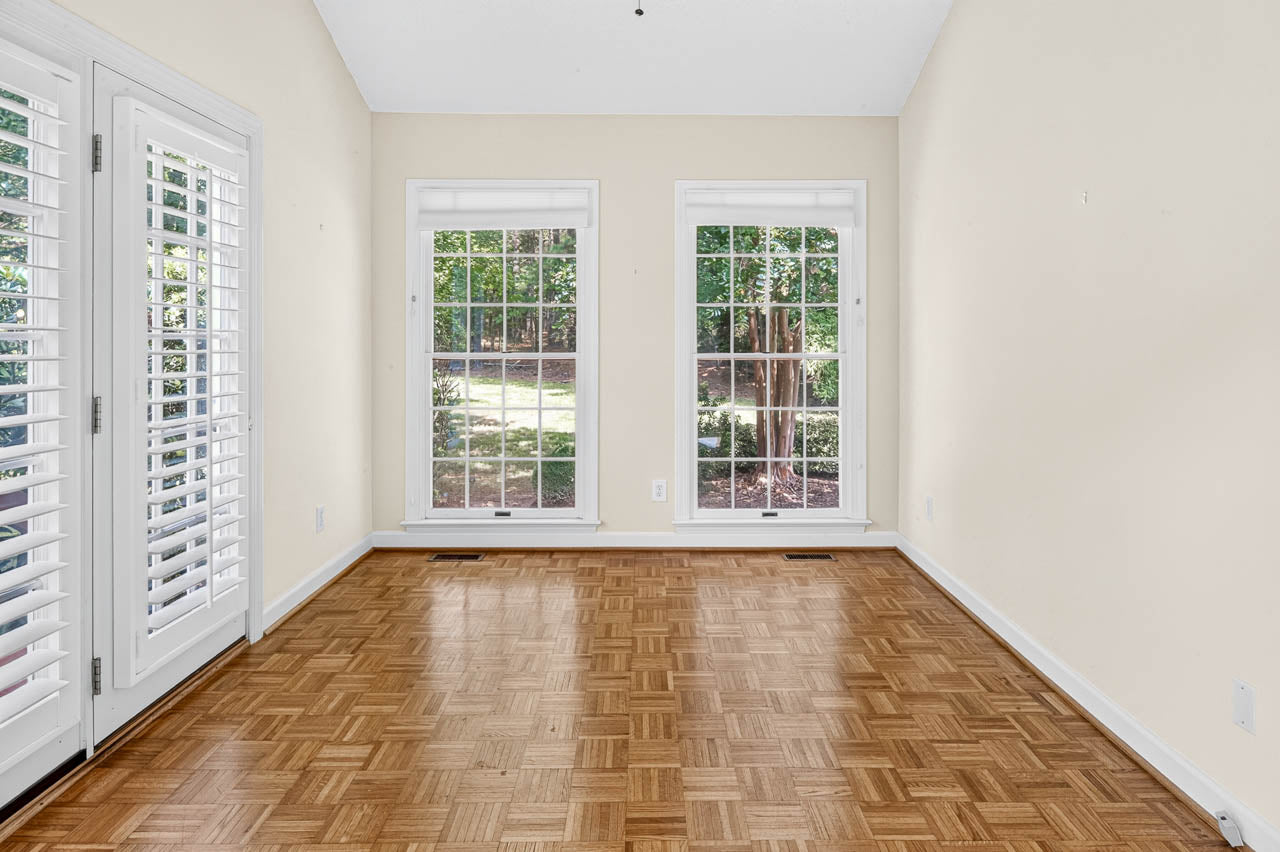 Empty room with light hardwood parquet flooring, cream-colored walls, and two large windows showing a green garden outside. Glass doors with white shutters are on the left. Bright natural light fills the space.