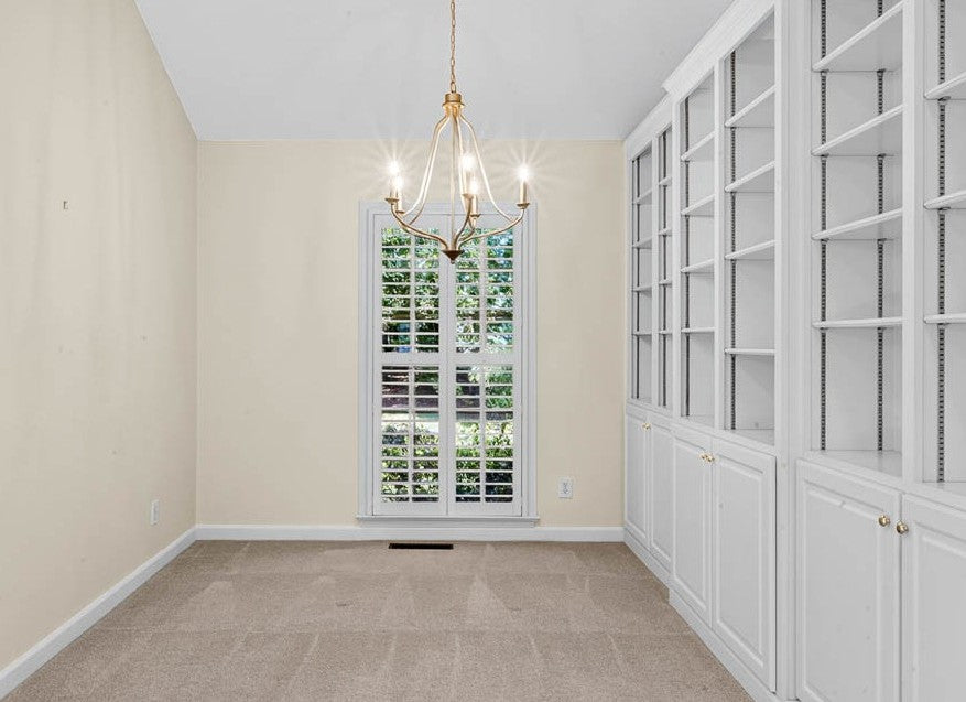 A bright room with beige walls, a large window with white shutters, an elegant gold chandelier, built-in white shelves and cabinets on the right, and light carpeted flooring.