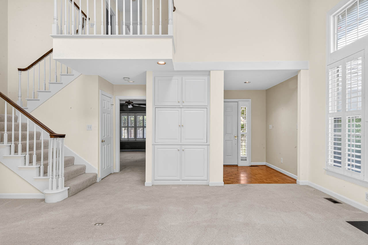 Bright living space with carpeted flooring, a staircase with a wooden handrail, built-in white cabinets, large windows with shutters, and an entryway with hardwood flooring.