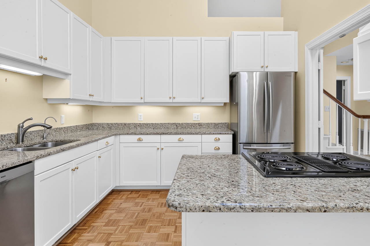 Modern kitchen with white cabinets, granite countertops, a stainless steel refrigerator, electric stove on an island, dishwasher, double sink, and light wood parquet flooring. Bright, clean, and spacious layout.
