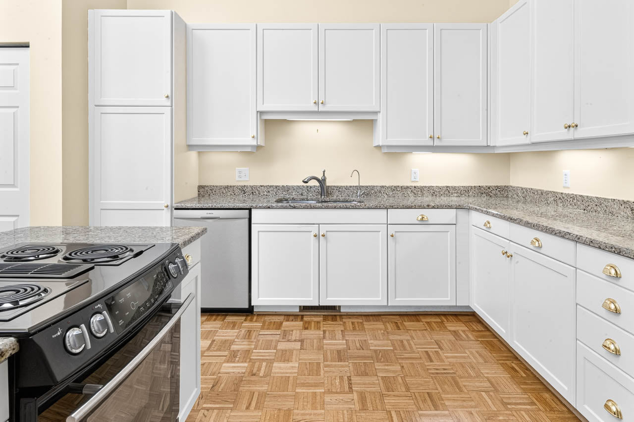 Modern kitchen with white cabinets, granite countertops, stainless steel appliances, electric stove, built-in oven, and parquet wood flooring. Sink and dishwasher are under cabinets, with ample storage space and neutral walls.