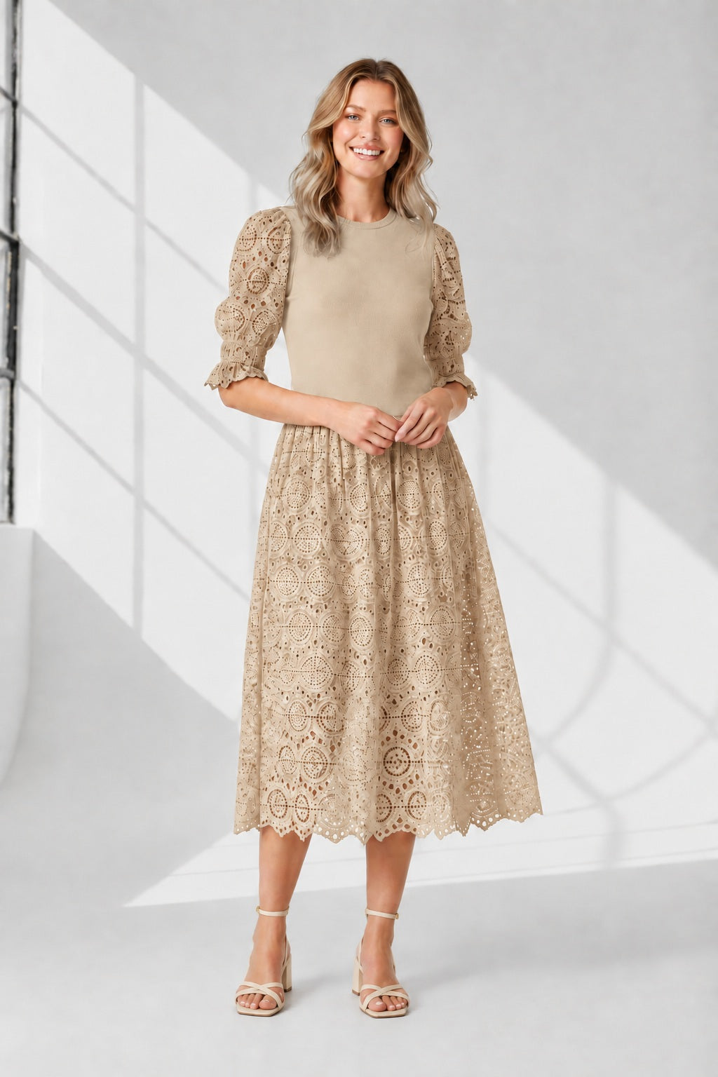 A woman stands against a white background wearing the SFIZIO - BRODERIE ANGLAISE MIDI DRESS by SFIZIO, featuring puffed sleeves and a lace skirt, paired with black strappy sandals.