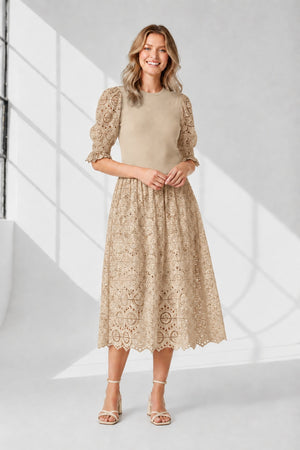 A woman stands against a white background wearing the SFIZIO - BRODERIE ANGLAISE MIDI DRESS by SFIZIO, featuring puffed sleeves and a lace skirt, paired with black strappy sandals.