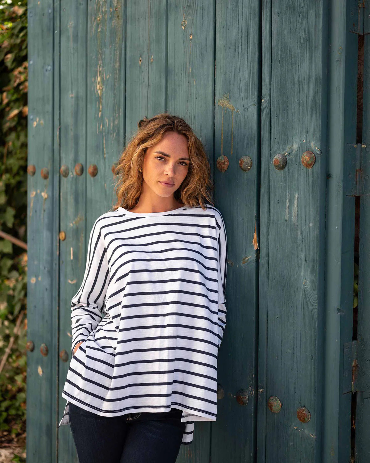 A woman with wavy brown hair models the MERSEA - CATALINA SLUB TEE—an oversized, long-sleeved white top with thin gray stripes—by MERSEA. She pairs it with blue jeans and stands against a plain, light background, hands behind her back.