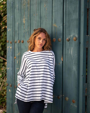 A woman with wavy brown hair models the MERSEA - CATALINA SLUB TEE—an oversized, long-sleeved white top with thin gray stripes—by MERSEA. She pairs it with blue jeans and stands against a plain, light background, hands behind her back.
