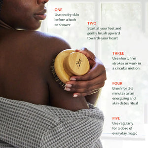 The KATE MCLEOD - THE DRY BRUSH, featuring natural bristles, is displayed on a beige towel atop marble, accented with an orange flower—evoking a relaxing dry brushing and lymphatic drainage ritual.