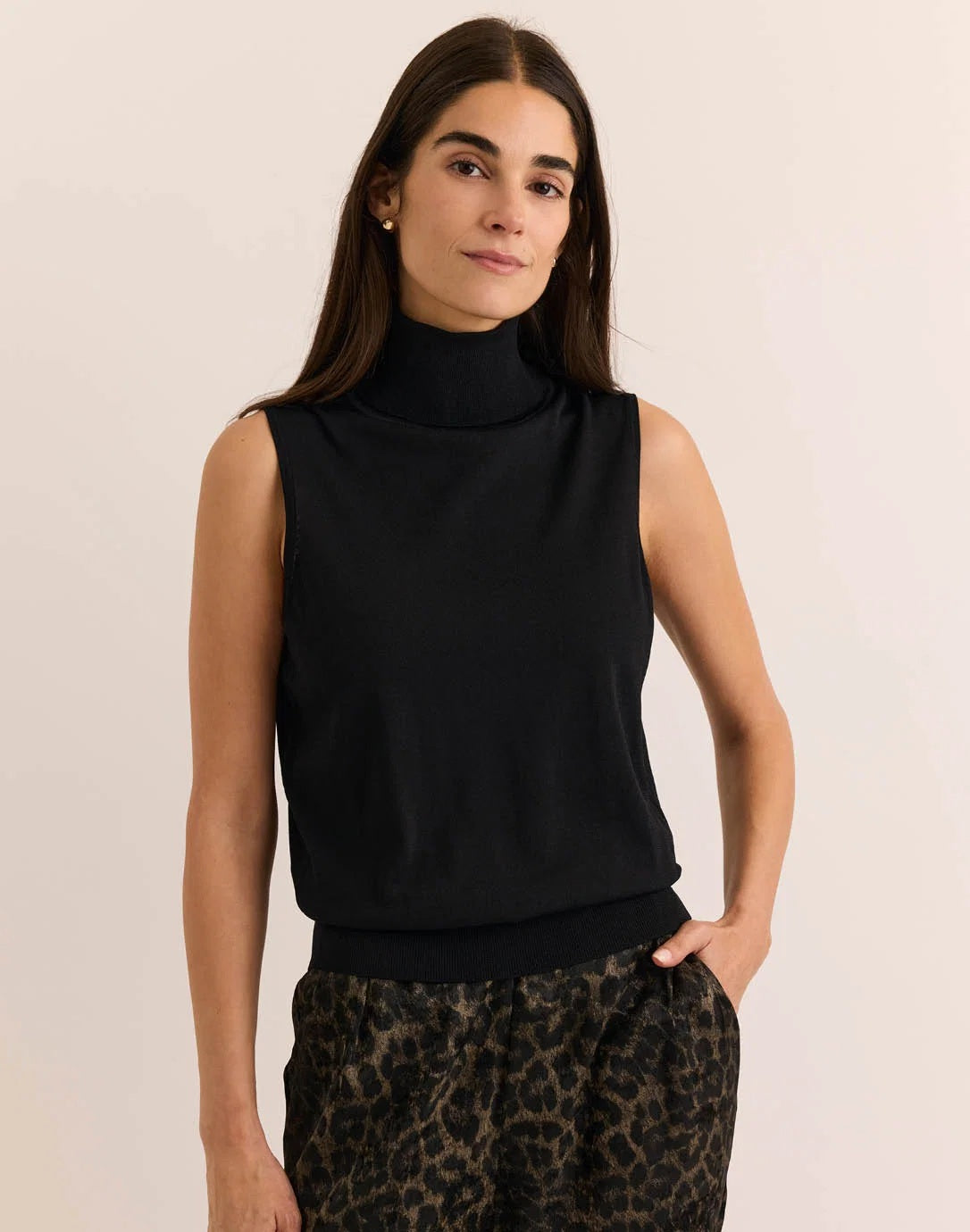 A woman with long brown hair wears the HINSON WU ASHLEY Sleeveless Turtleneck Knit Tank in black, paired with leopard print pants, standing against a plain light background with one hand on her hip.