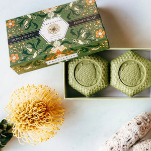 A beautifully packaged BAUDELAIRE Calendula Honey Soap Gift Box includes two 3.5 oz hexagonal green bars with an embossed beehive motif, partially displayed from a decorative floral box labeled “Calendula.” This elegant BAUDELAIRE gift set features triple-milled soap for a luxurious and gentle cleansing experience.