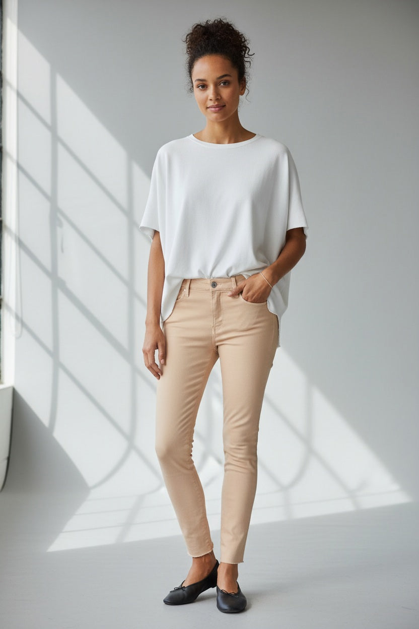 A person stands in a kitchen, showing only their lower half, wearing FRANK &amp; EILEEN’s SLIGO SKINNY JEAN in light beige with a white top and black flats, set against a stove, countertop, and patterned rug.