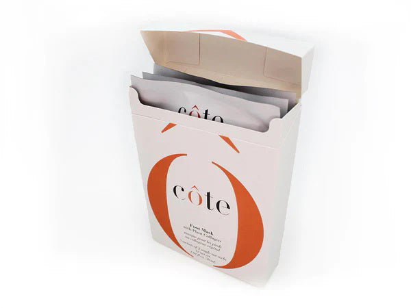 A white rectangular box partially open at the top, revealing multiple sheet masks inside, along with a special foot mask for age-defying hydration. The box is labeled with the brand name "COTE" and adorned with orange design accents. It contains "COTE - FOOT MASK WITH COLLAGEN 3 PACK.