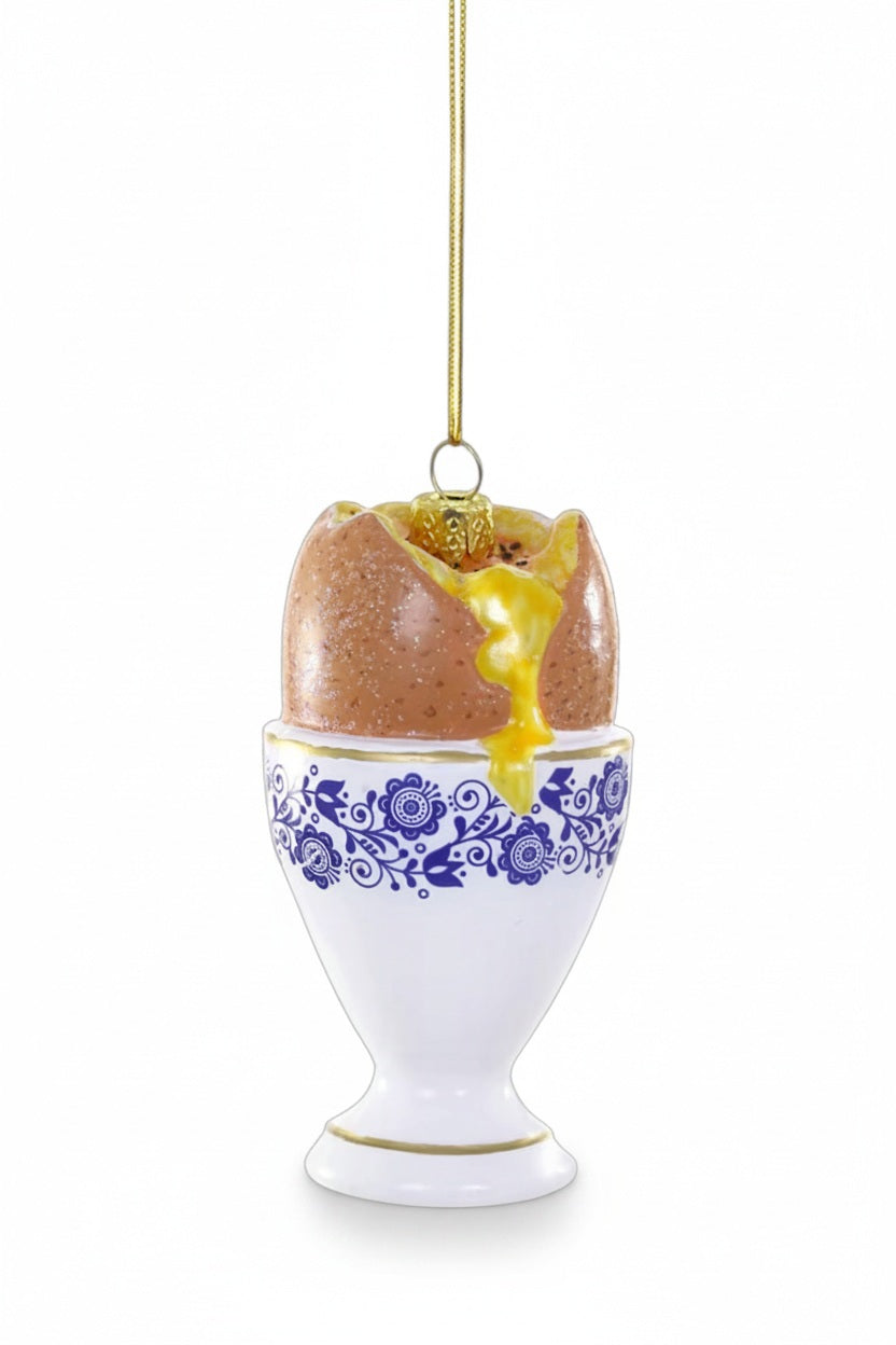 Cody Foster EGG IN A CUP ORNAMENT, a 3.5-inch glass Christmas ornament shaped like a cracked, soft-boiled egg in a blue and white floral patterned egg cup.