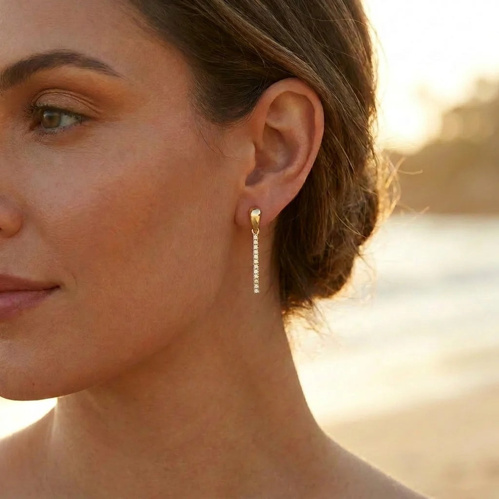 Introducing the ELLA STEIN - DROP THE MIC EARRINGS by ELLA STEIN: a pair of gold plated drop earrings featuring a sleek, vertical bar design, each adorned with a row of shimmering white stones.